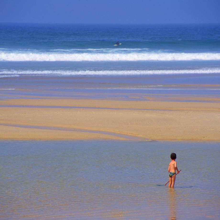 Enfant sous le soleil à la plage, au loin un surfeur dans les vagues