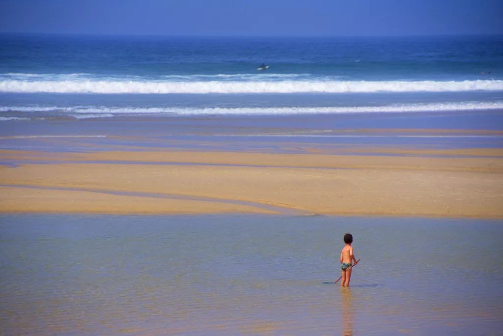 Enfant sous le soleil du matin à la plage, au loin un surfeur dans les vagues